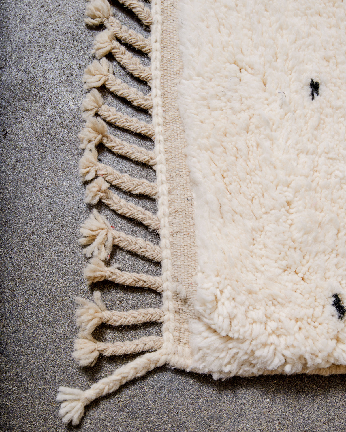 Corner of cream Beniourain wool rug with a tightly woven edge and natural wool tassels laying on concrete floor.